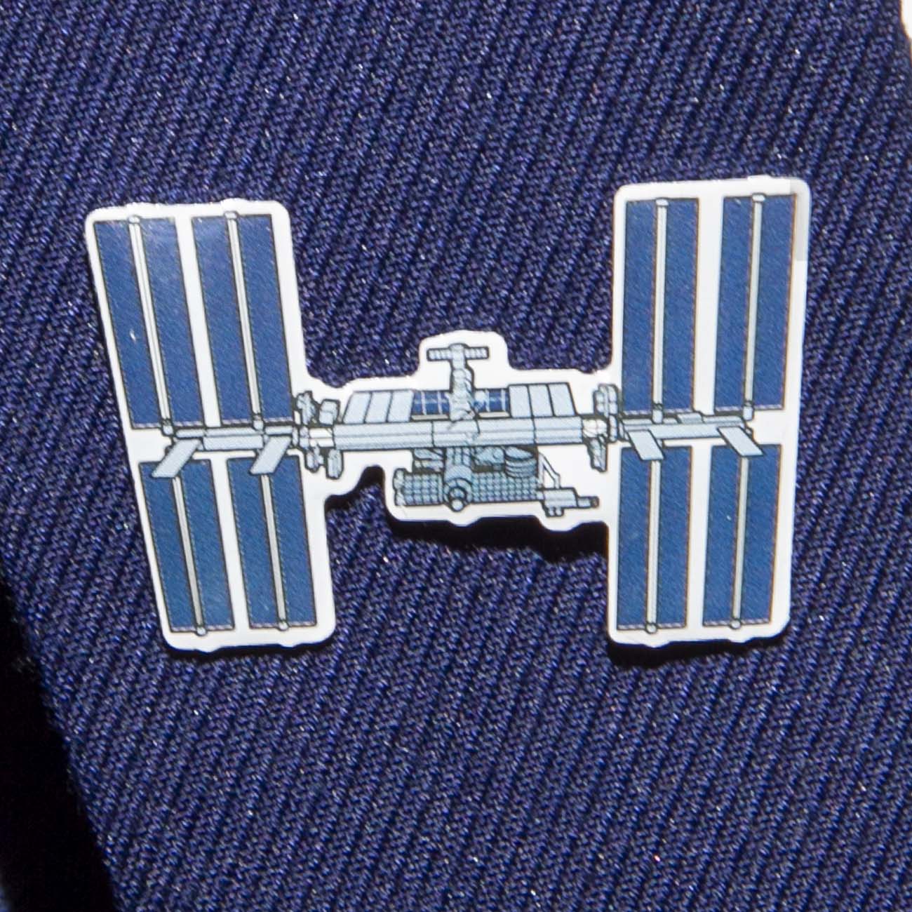 A close-up of the Boeing Illustrated ISS Lapel Pin by The Boeing Store shows a silver tone pin shaped like the International Space Station, with solar panels and modules in an epoxy finish, attached to dark navy textured fabric.